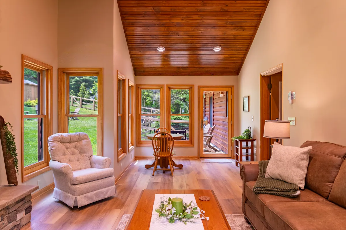 A cozy armchair and wooden dining chair sit near a coffee table adorned with tasteful decor, next to large windows offering scenic views. Sunlight streams into the room, blending comfort with a sense of openness to the outdoors.