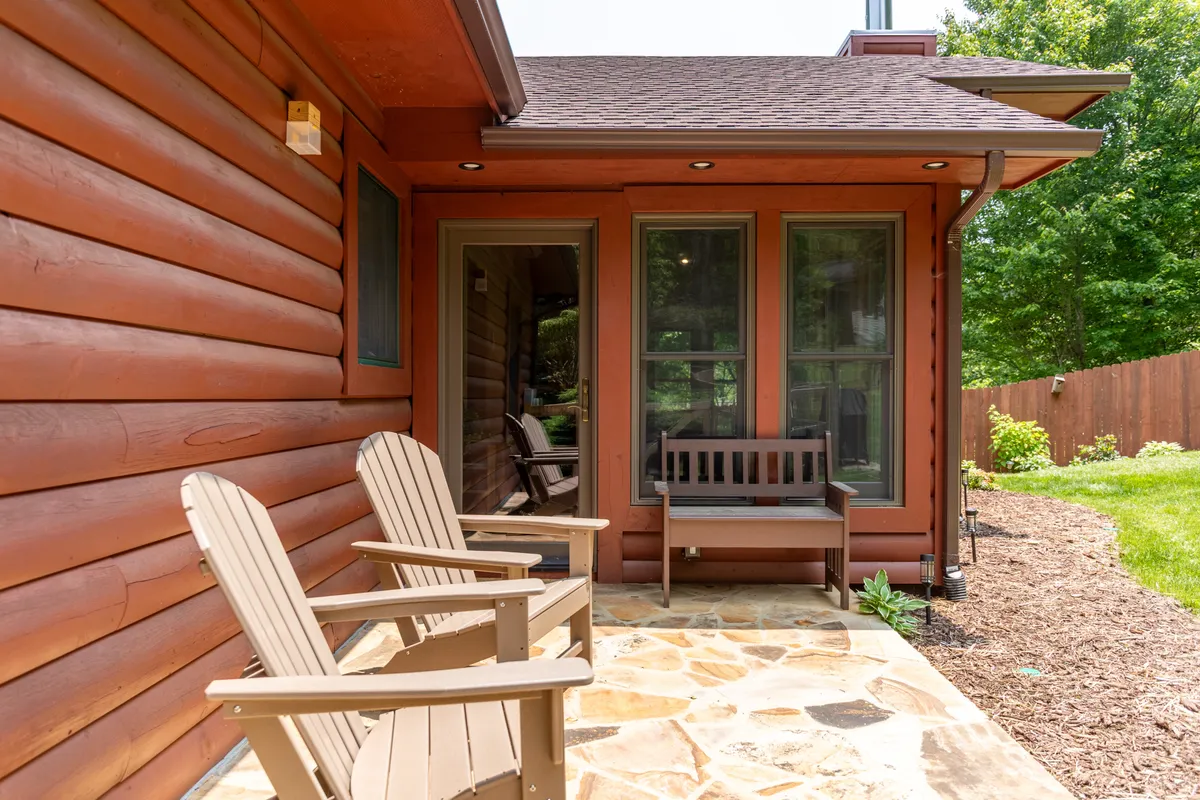 A stone patio featuring wooden chairs and benches is surrounded by tranquil greenery. The setup suggests a space for families to relax, socialize, and enjoy nature, evoking the mood of evenings by the firepit and group activities.