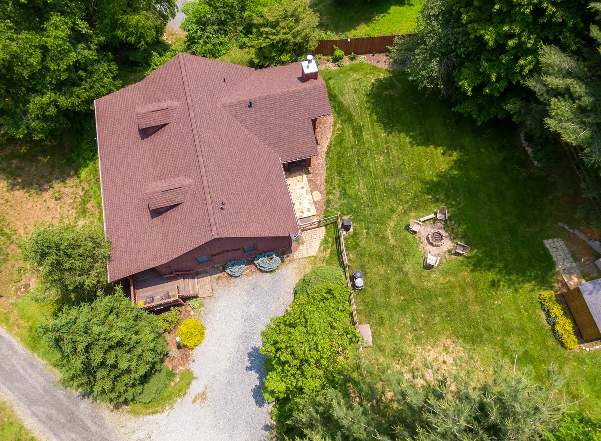 Here, the house is visible with its green lawn and a stone fire pit set in the center, surrounded by trees and open sky. The setting is open yet enclosed by nature, perfect for outdoor games and gatherings. The view captures the expanse of the yard and the versatility of the space for different activities.