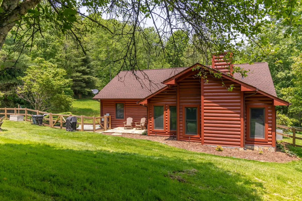 This image showcases a beautiful log cabin structure with an expansive green lawn and a visible fenced yard. The patio area is furnished with chairs, and a barbecue grill is set up, creating a welcoming outdoor space. The fence is clearly visible, providing a sense of security and privacy. Lush greenery surrounds the area, enhancing the mountain serenity.