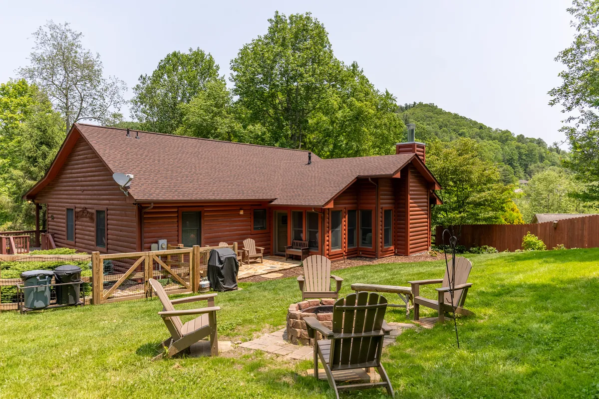 The image showcases a classic log cabin nestled among lush greenery, with a spacious, manicured yard bordered by mature trees. In the foreground, a cozy fire pit is surrounded by a circle of inviting Adirondack chairs, perfect for gathering. The golden glow of sunset bathes the scene, highlighting the warm, welcoming ambiance of the outdoor space.