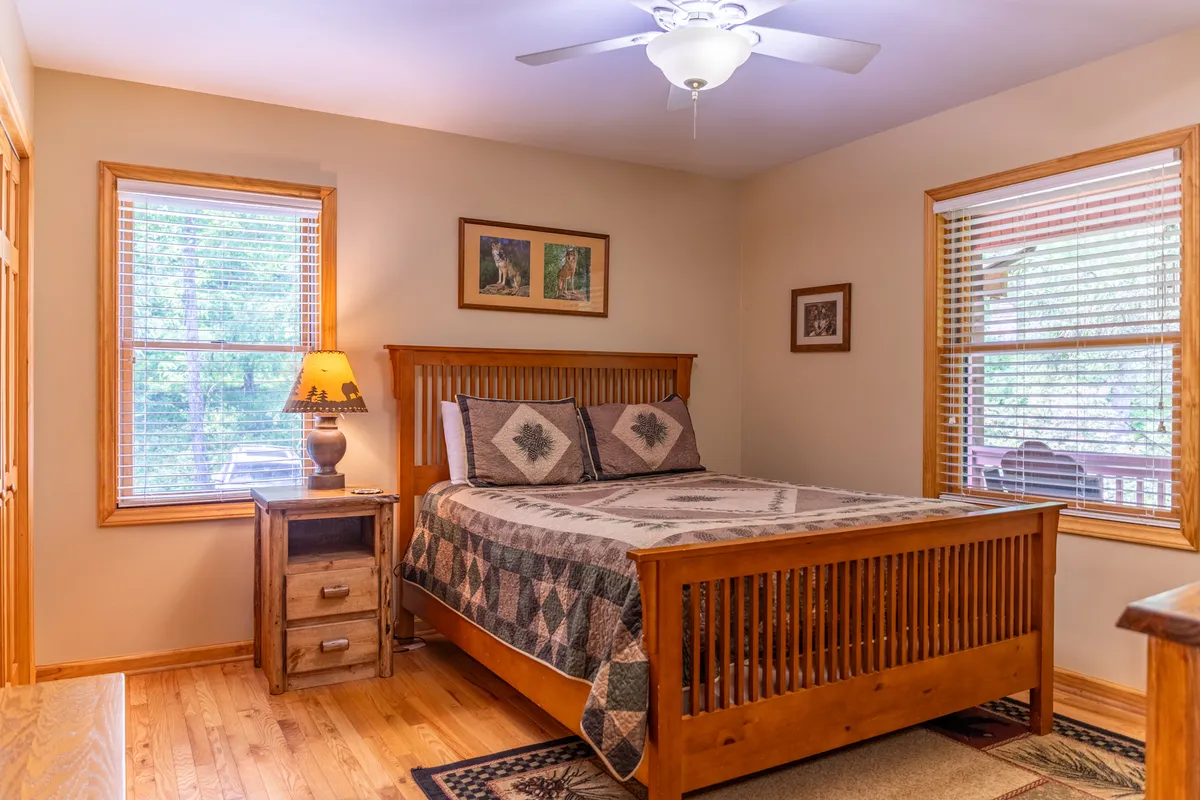A warmly lit bedroom featuring a comfortable bed, nightstand with a lamp, windows letting in natural light, and artwork on the wall. The space feels inviting and calm, ideal for relaxation or waking up to peaceful views.