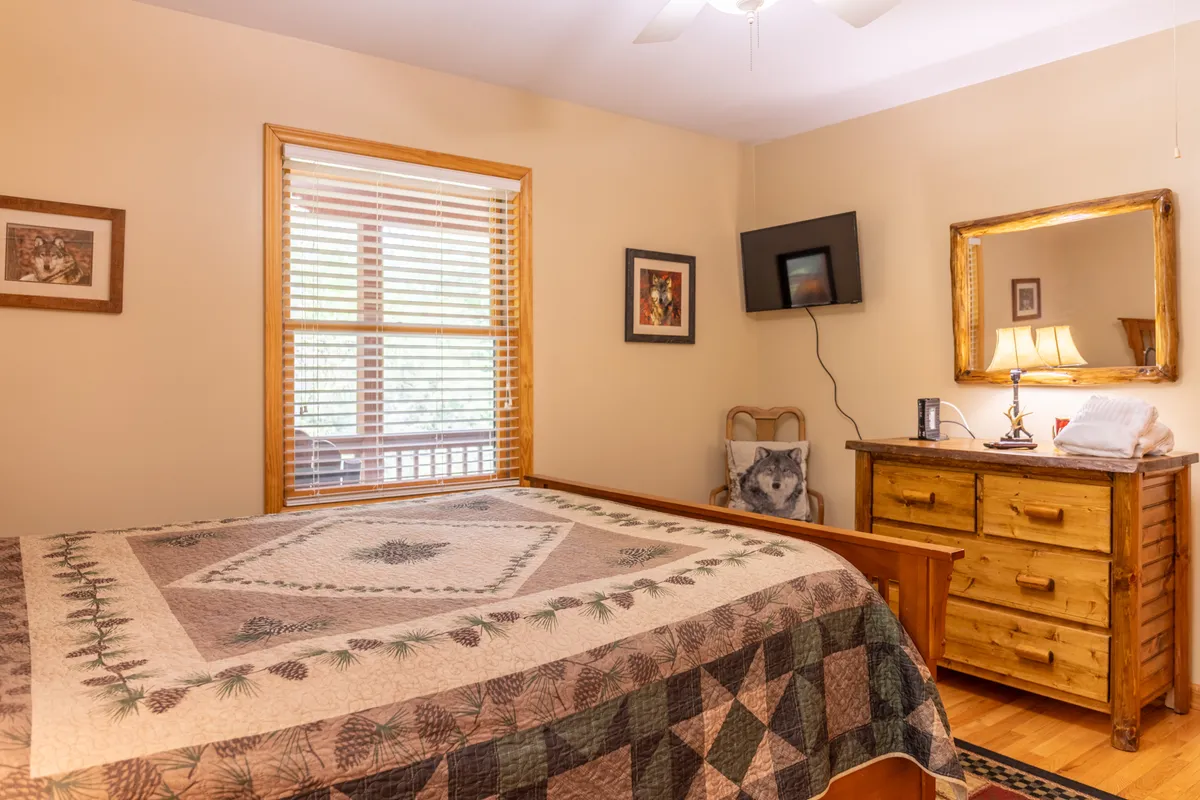 A tranquil bedroom scene with a quilted bedspread, wooden dresser, framed artwork, and a window with blinds. The atmosphere is peaceful, ideal for enjoying quiet moments or a morning coffee by the window.