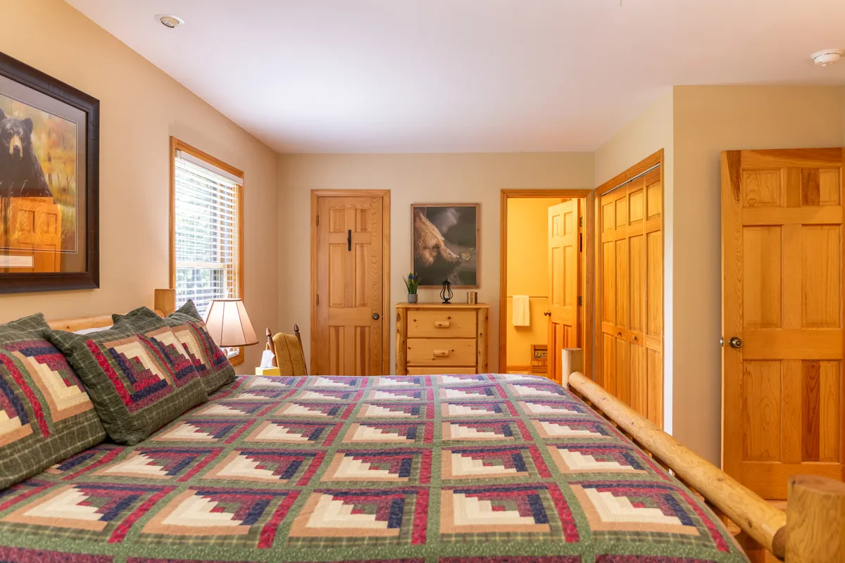 A serene bedroom featuring a bed with a patterned quilt, wooden furniture, and artwork of a bear on the wall. Soft natural light filters through the window, creating a peaceful and cozy atmosphere.