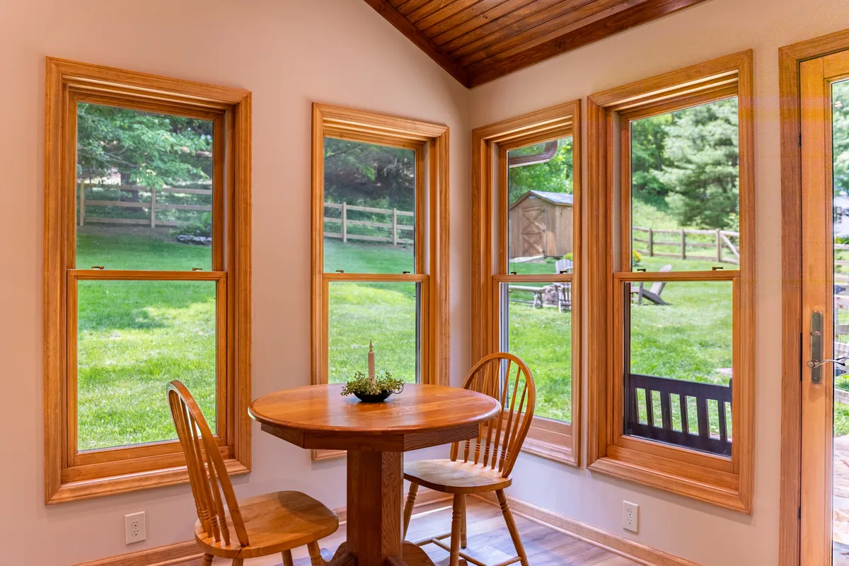 This image features a round wooden table and four chairs set beside large, light-filled windows overlooking a lush green landscape. The natural light pouring in and the open view create an inviting, sun-drenched corner ideal for morning coffee or quiet reflection.