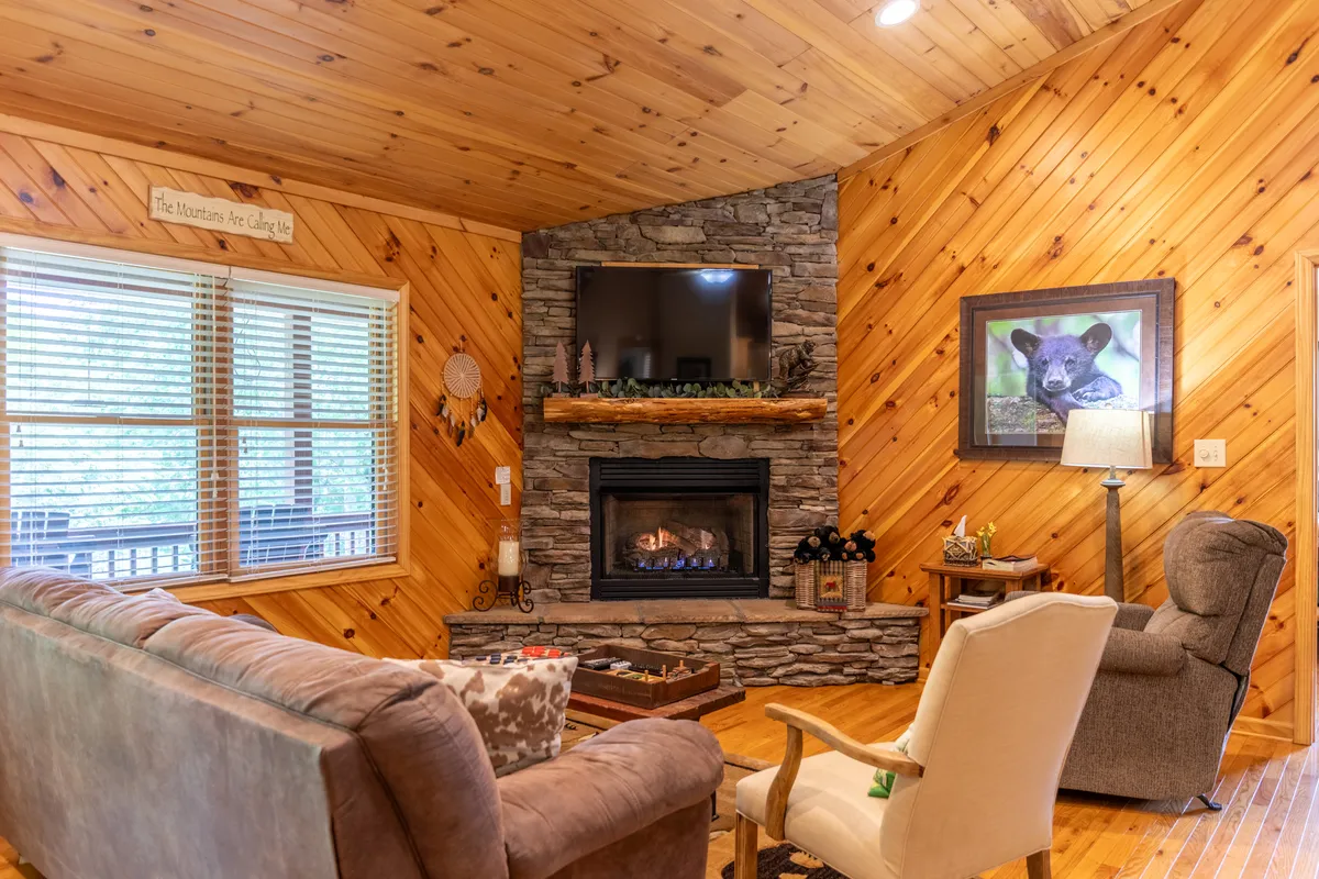 The living room features a fireplace, sofa, armchair, TV, and wall art. Windows offer views of nature, and the space is styled to invite socializing and relaxation, suggesting a comfortable environment for both work and leisure.