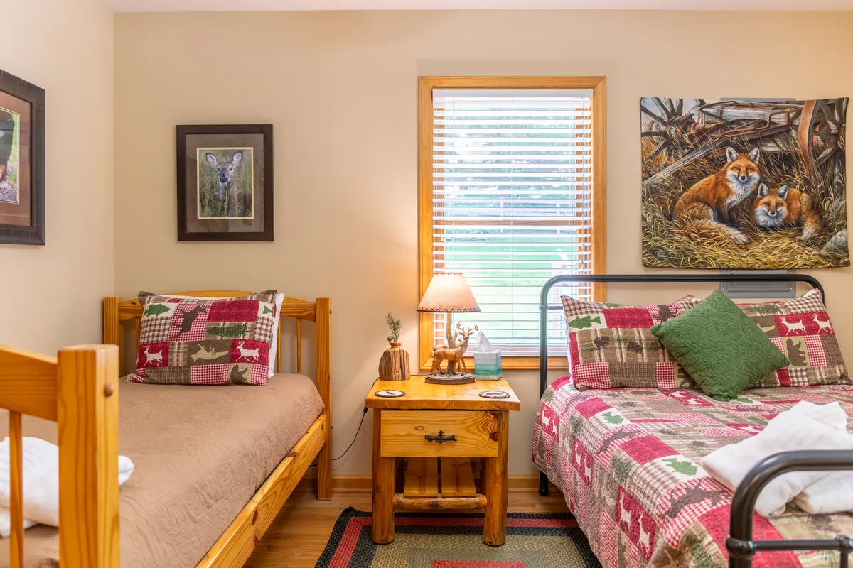 A cozy bedroom with wooden beds, plaid bedding, a nightstand lamp, fox painting, and framed deer art. The large window allows natural light to stream in, hinting at mountain views and the surrounding landscape. The decor is nature-inspired and engaging, complimenting the peaceful mountain setting referenced in the section.