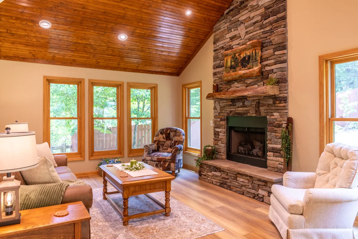 This image displays a spacious great room featuring a beautiful stone fireplace, wood-paneled ceiling, comfortable seating, and large windows that fill the space with natural light. The inviting atmosphere is perfect for gathering, reading, or relaxing by the fire.