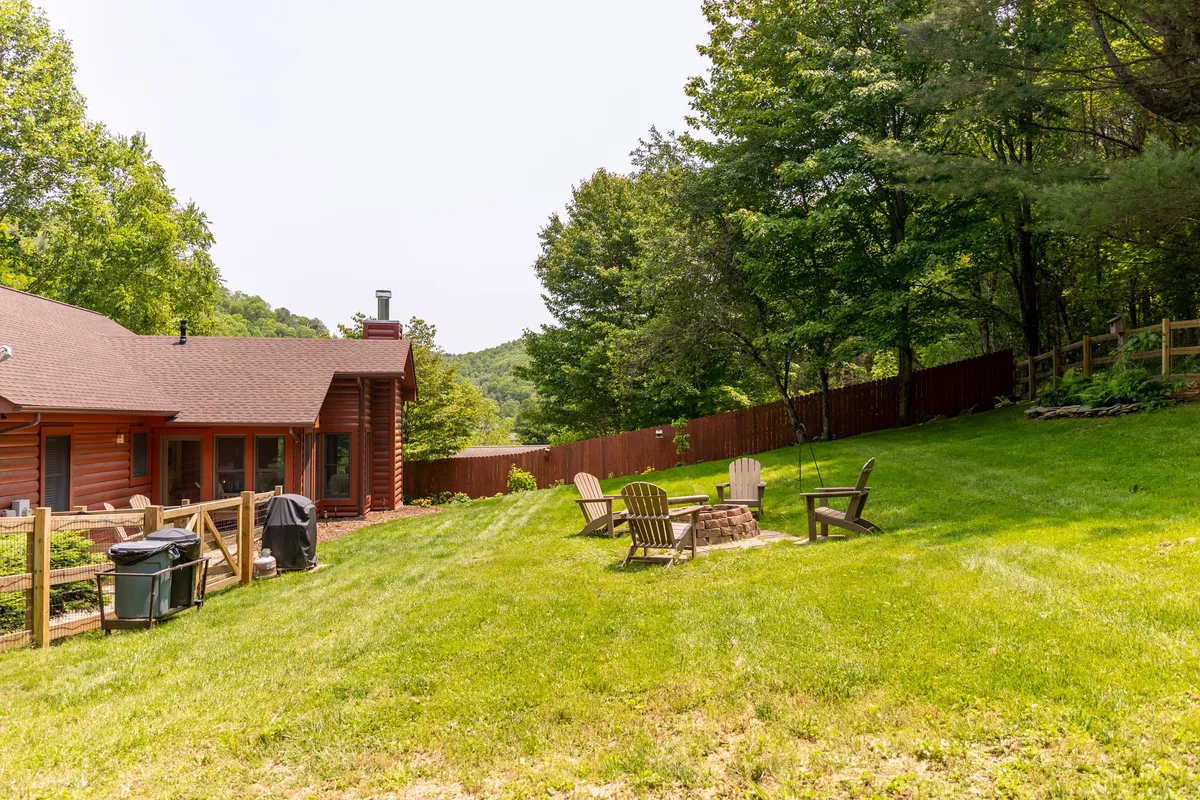 This inviting exterior shot features a log cabin with a freshly mowed grassy backyard, a prominent stone fire pit surrounded by Adirondack chairs, and lush trees for privacy. The area is clearly designed for gatherings, relaxation, and making memories under the open sky.